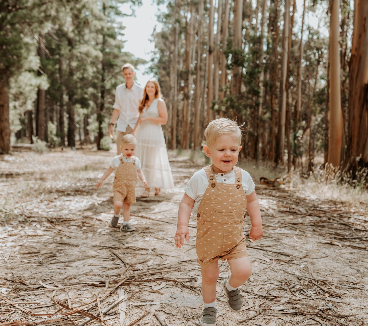 Playful toddlers in tan overalls explore a sunlit forest path together.