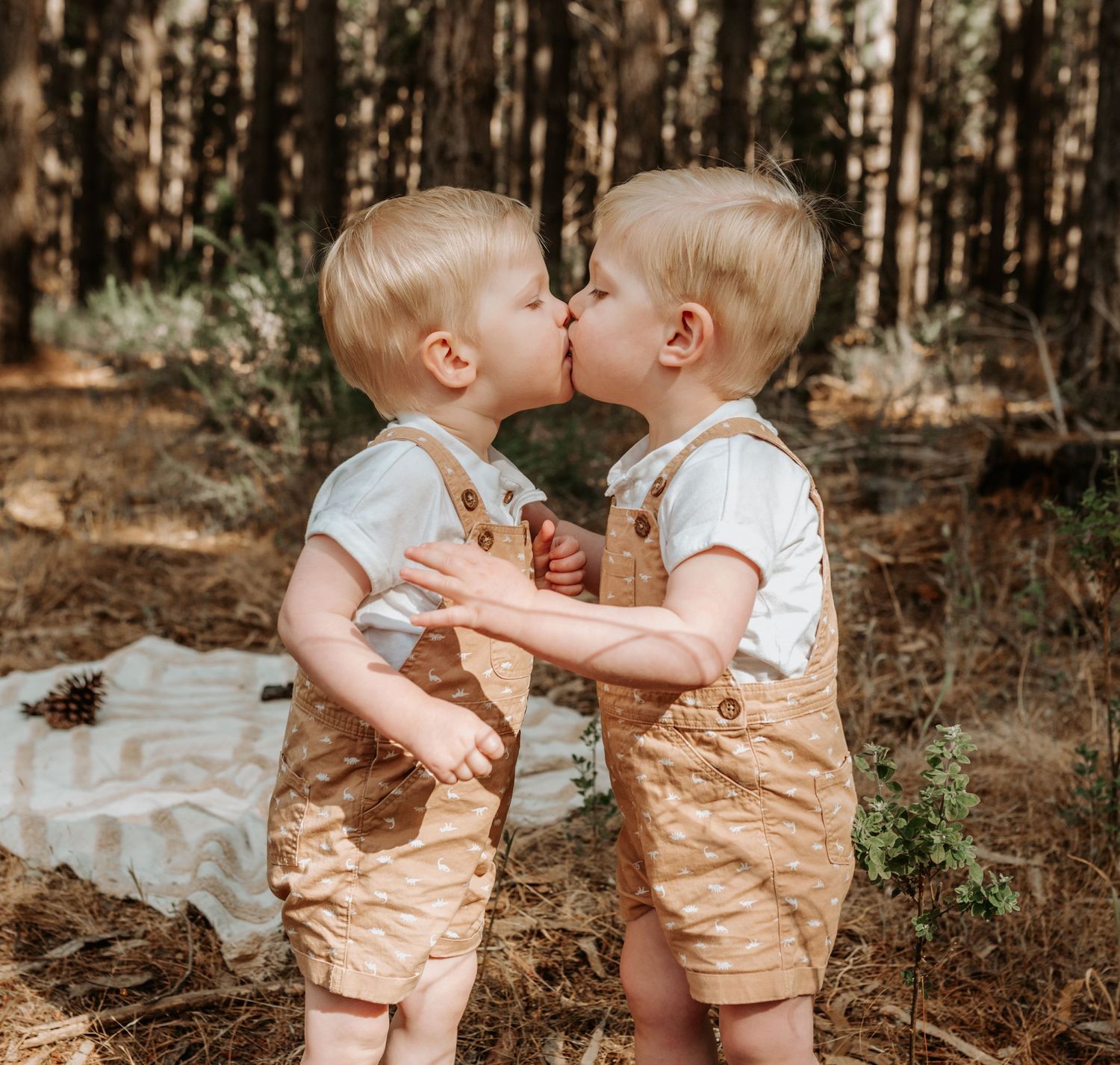 Two small children in matching overalls share a sweet moment in a wooded setting.