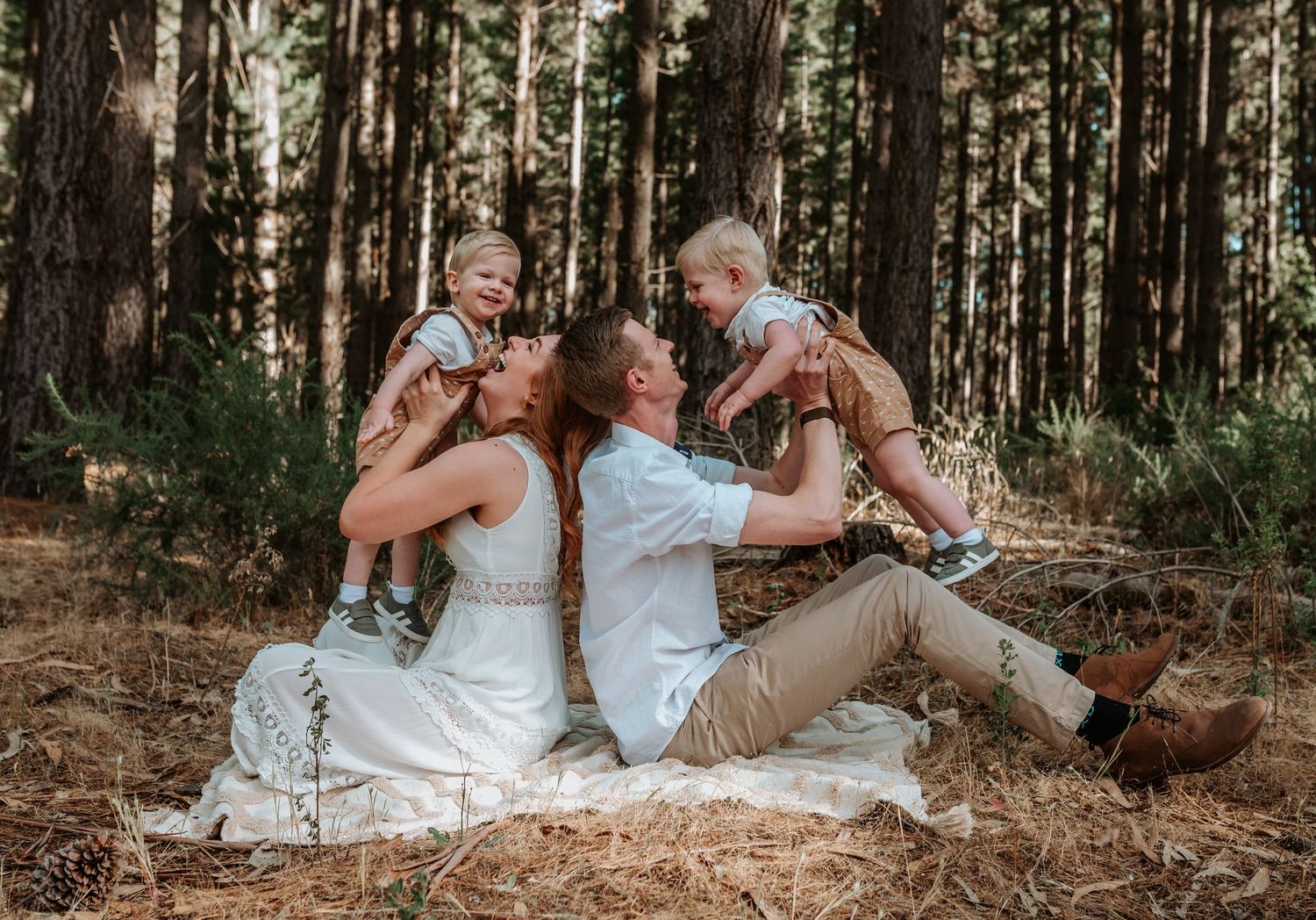 A couple in wedding attire sits together on a forest floor surrounded by tall pine trees.