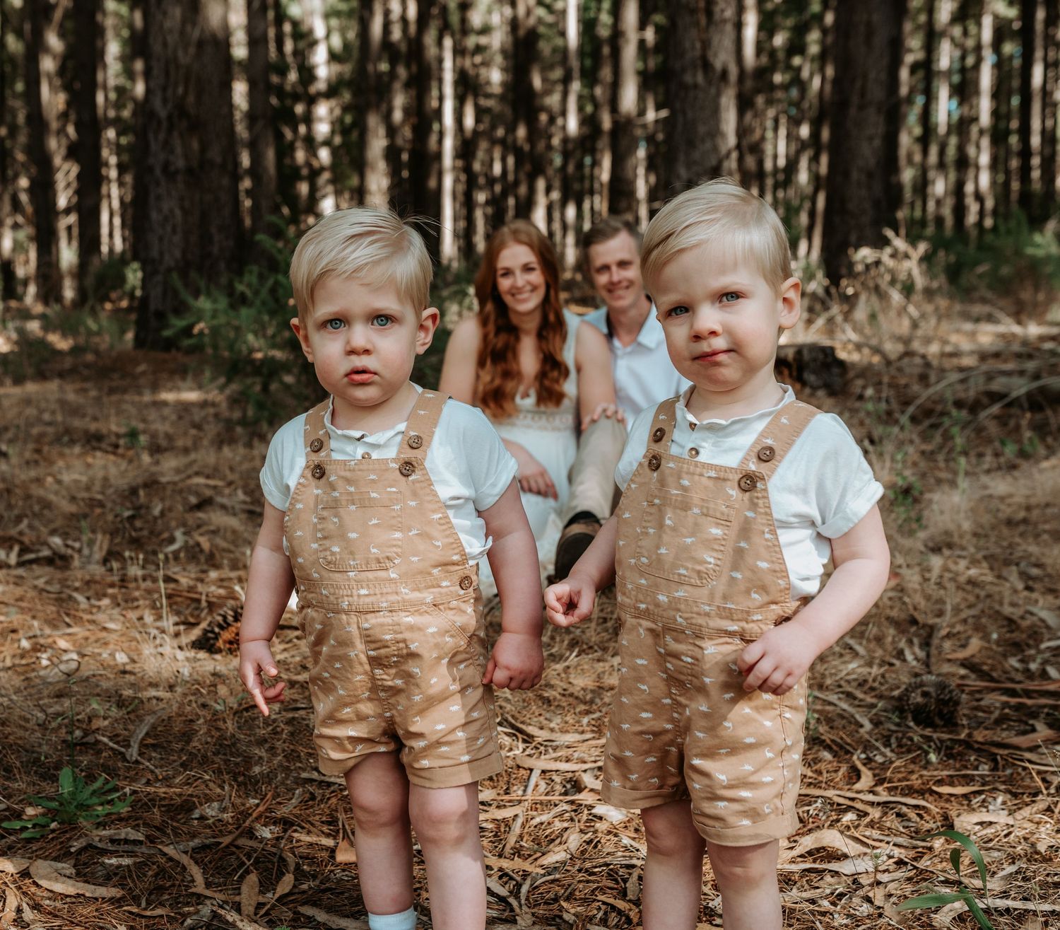 Two children in coordinating outfits stand together among tall pine trees.