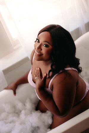 A smiling woman in pink lingerie relaxes in a bubble bath, looking over her shoulder in a bright, airy room.