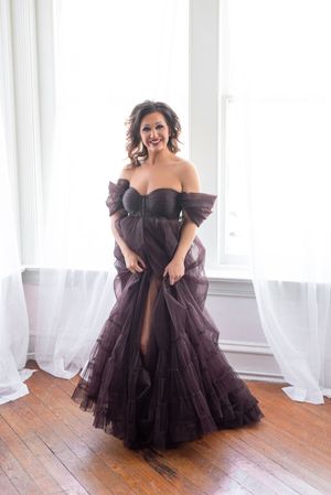 Woman in elegant off-shoulder dark plum tulle gown posing gracefully in bright windowed room with hardwood floors.