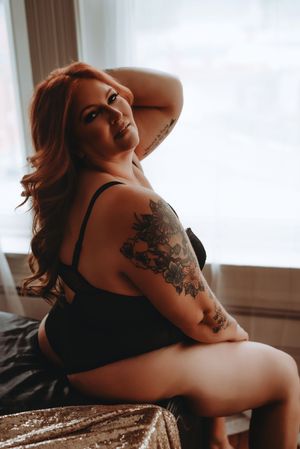 A redhead woman with floral arm tattoos poses in black lingerie on a bed near a bright window.