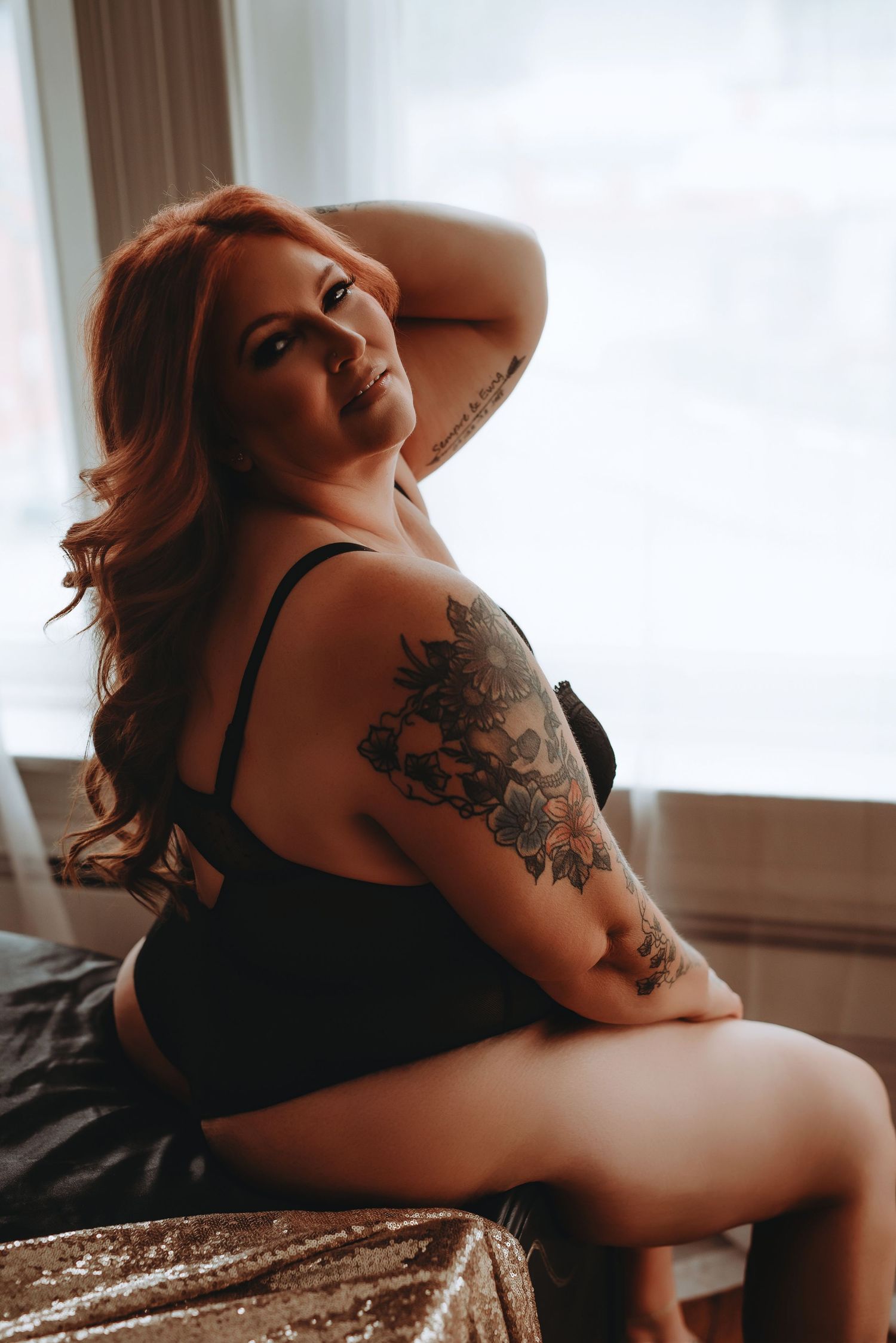 A redhead woman with floral arm tattoos poses in black lingerie on a bed near a bright window.