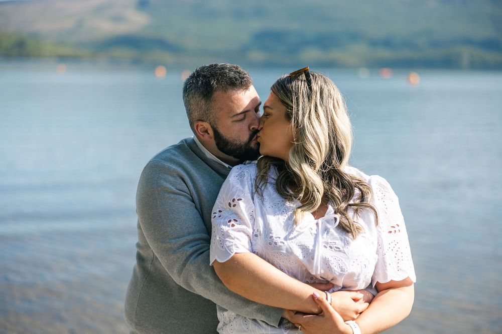A couple embraces by a scenic lake with mountains in the background.