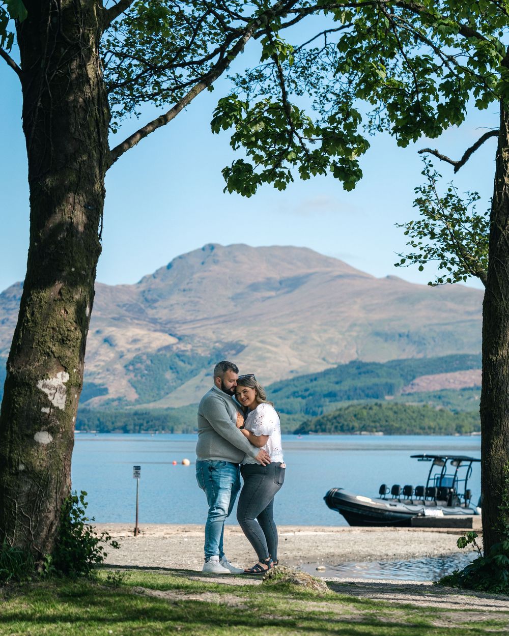 A couple shares a playful moment by a serene lake with mountains and trees framing the scene.