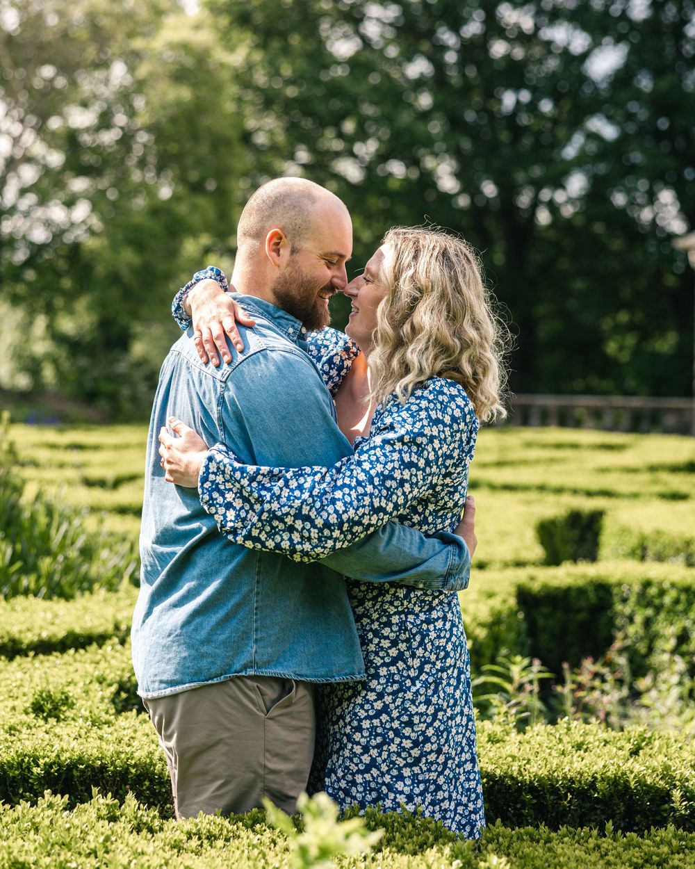 A couple embraces in a sunny garden setting with green hedges in the background.