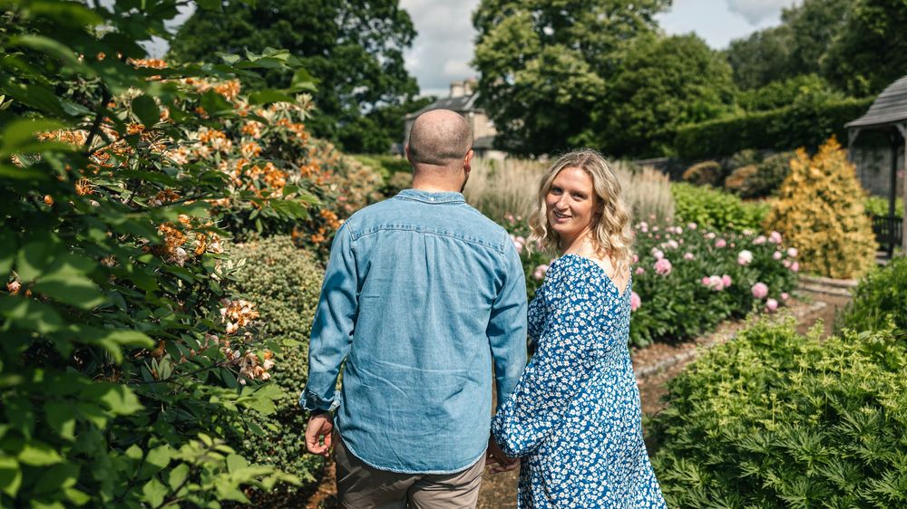 A couple shares a romantic moment in a manicured garden with trees and shrubs.