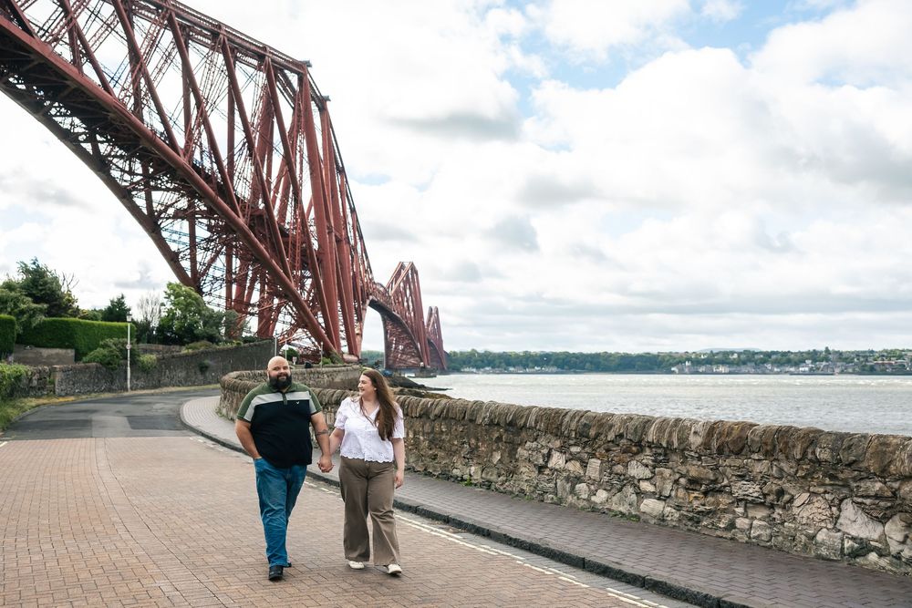 A couple shares a romantic moment under the iconic Forth Bridge in Scotland on a cloudy day.