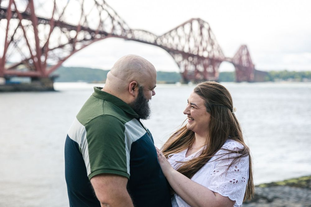 Romantic couple embracing near the red steel beams of the Forth Bridge in Scotland.