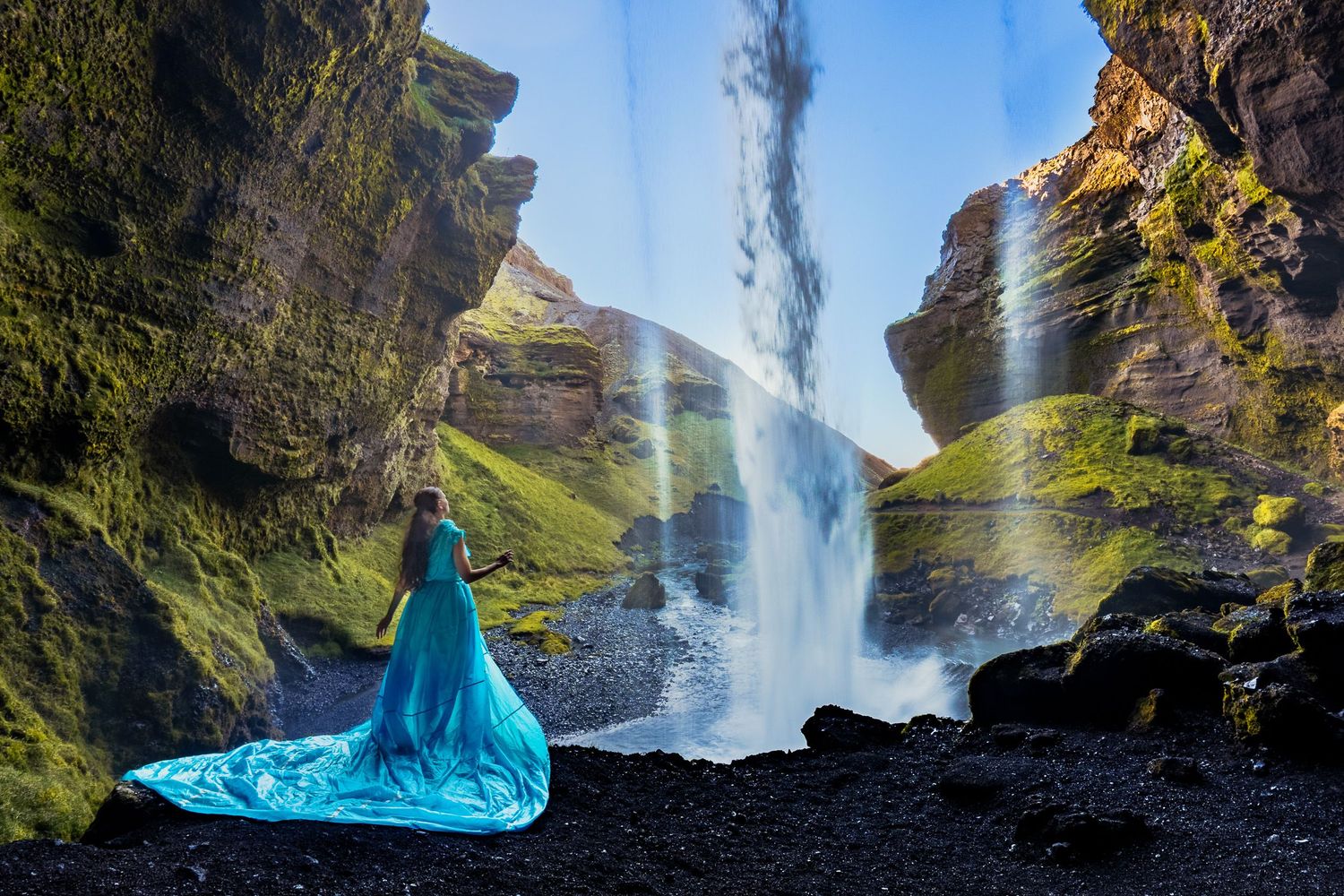 Woman in flowing turquoise gown stands before a majestic waterfall cascading down moss-covered rocky cliffs.