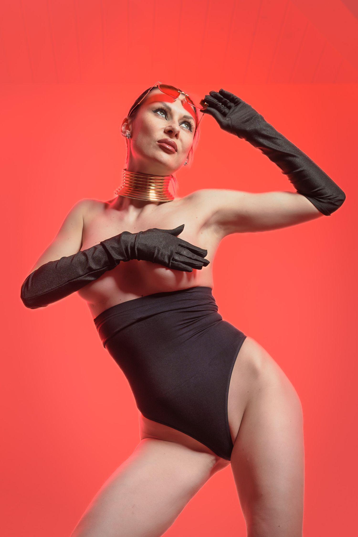 Woman in black high-waist bikini and long gloves posing dramatically against vibrant red background with gold neck cuff.
