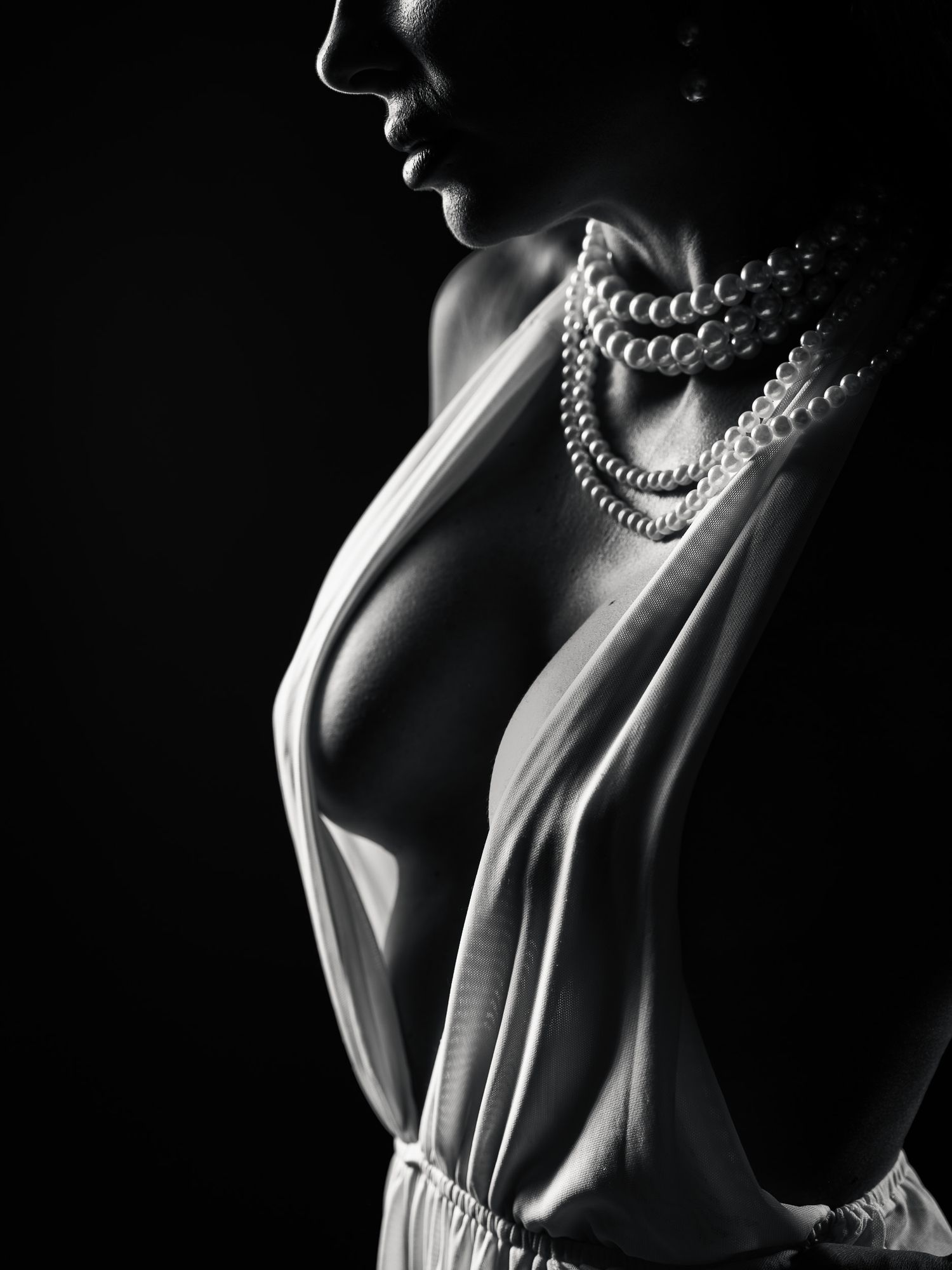 Black and white portrait of a woman in a low-cut white dress wearing layered pearl necklaces against a dark background.