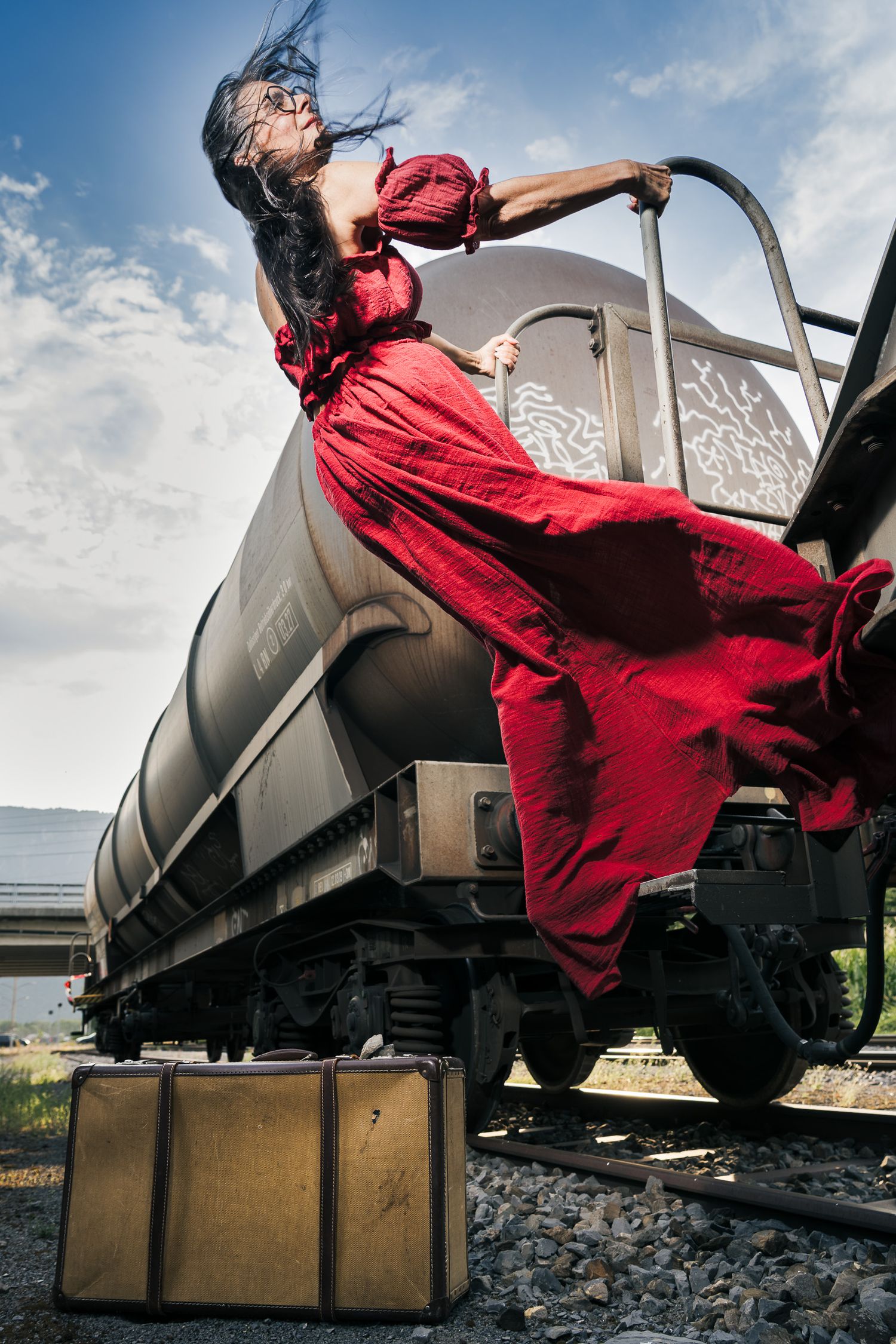 Woman in flowing red dress clinging to a moving train car on railroad tracks with a vintage suitcase below.