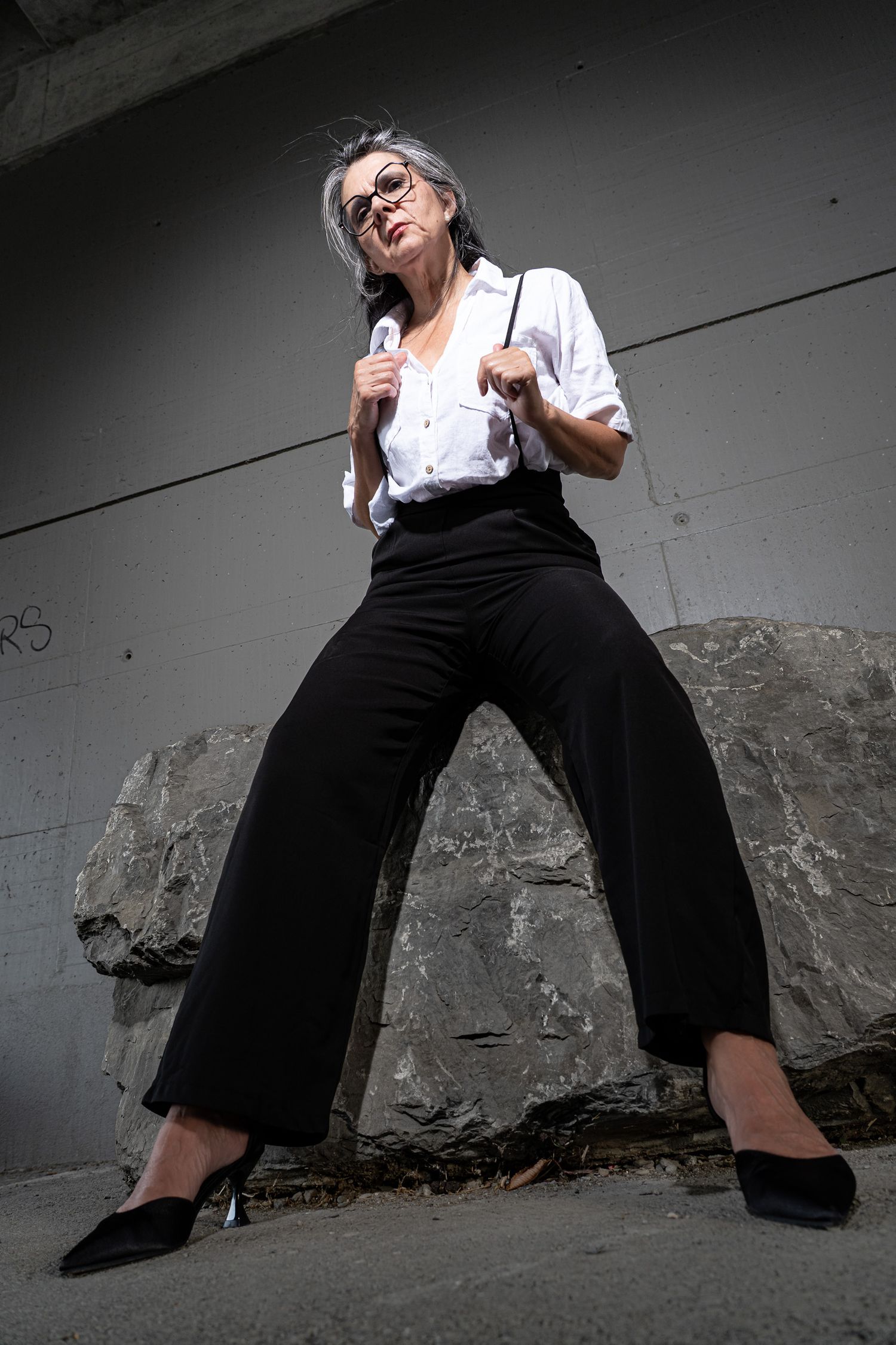 A confident older woman with gray hair poses in black wide-leg pants, white shirt, suspenders, and heels outdoors.