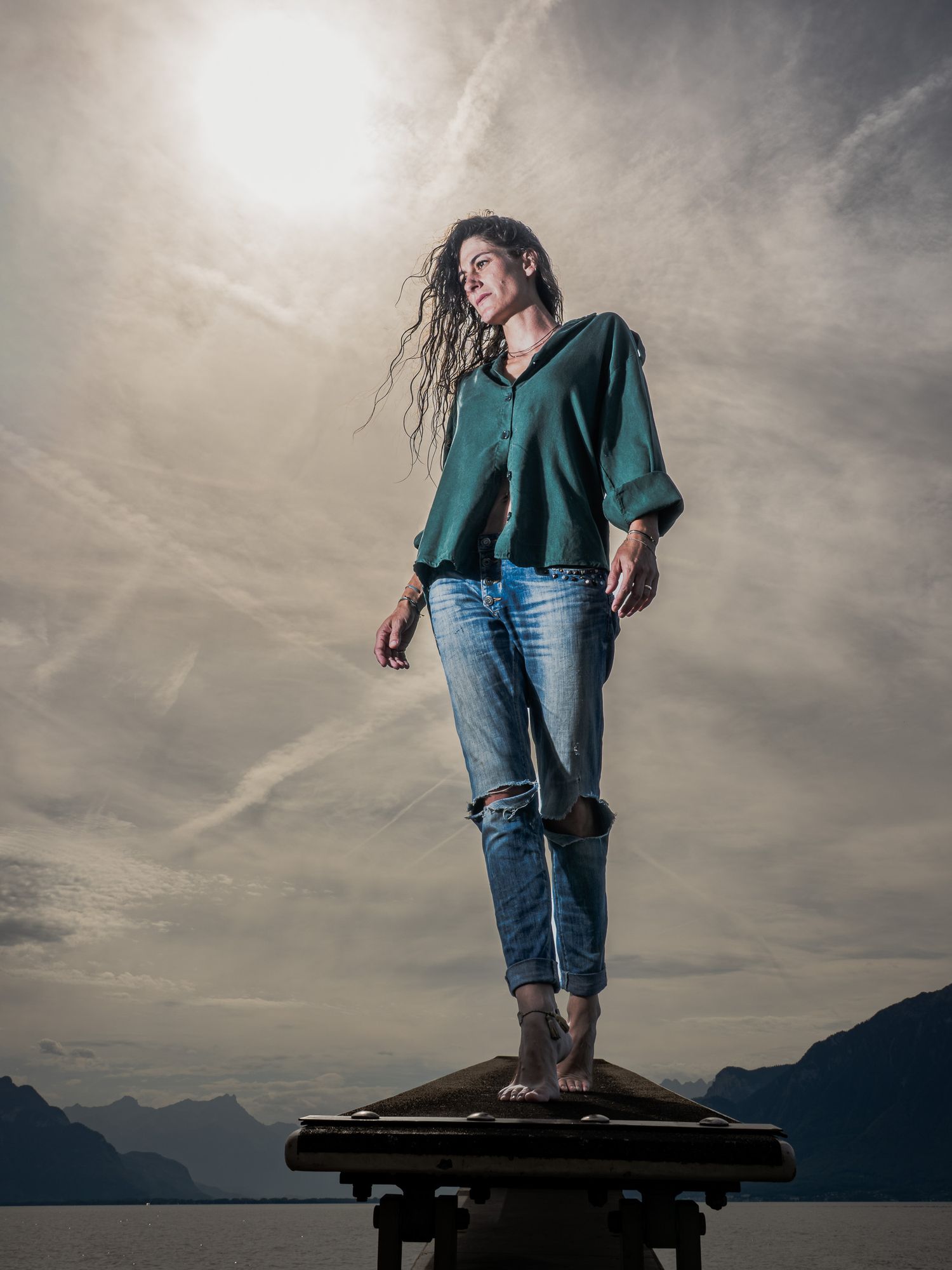 Woman in green blouse and ripped jeans stands barefoot on platform overlooking mountain lake under dramatic cloudy sky.