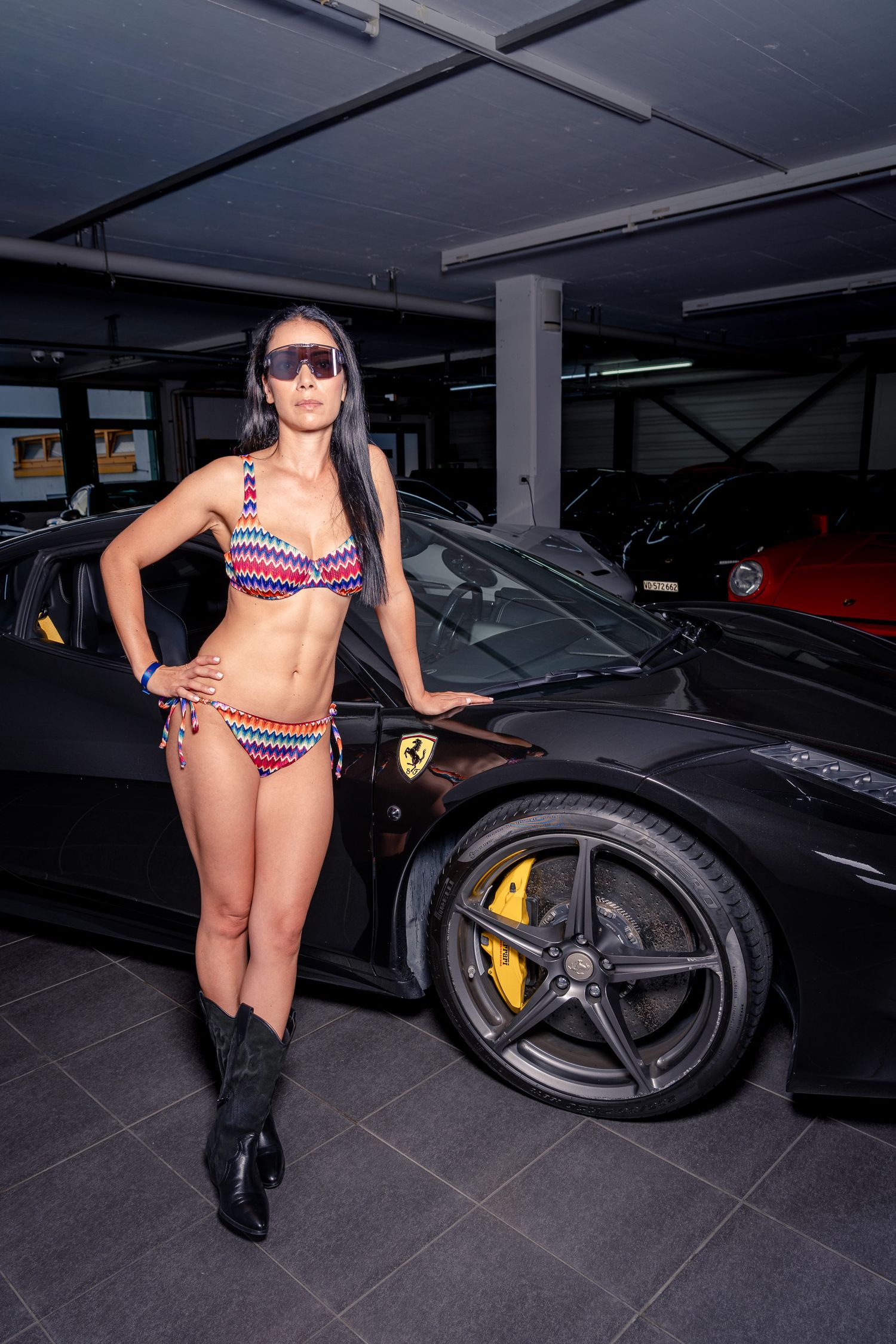 Woman in colorful bikini and cowboy boots posing beside a black Ferrari in an underground garage.