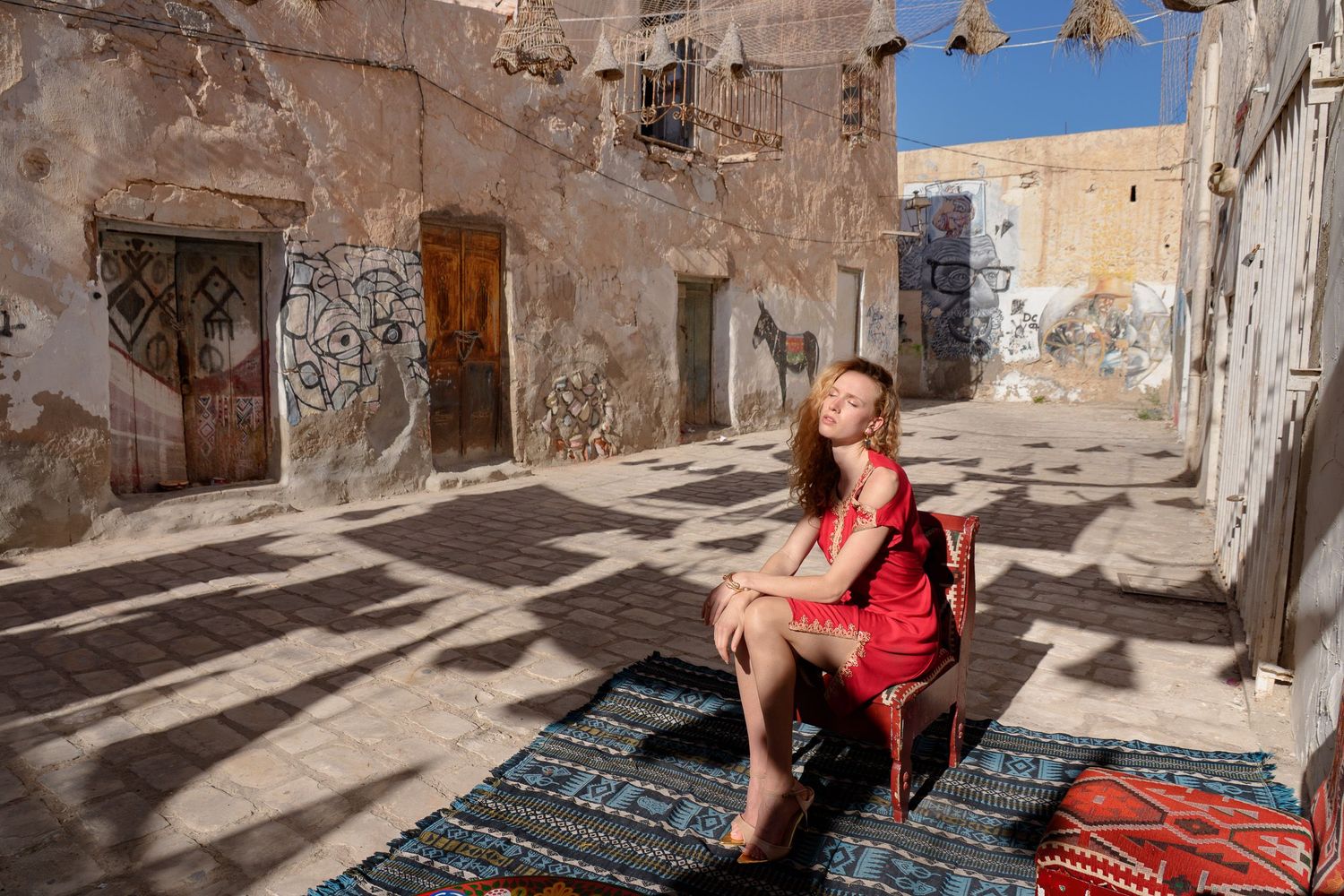 Woman in red dress sitting on chair in sunlit Moroccan courtyard with ornate doors and colorful rugs.