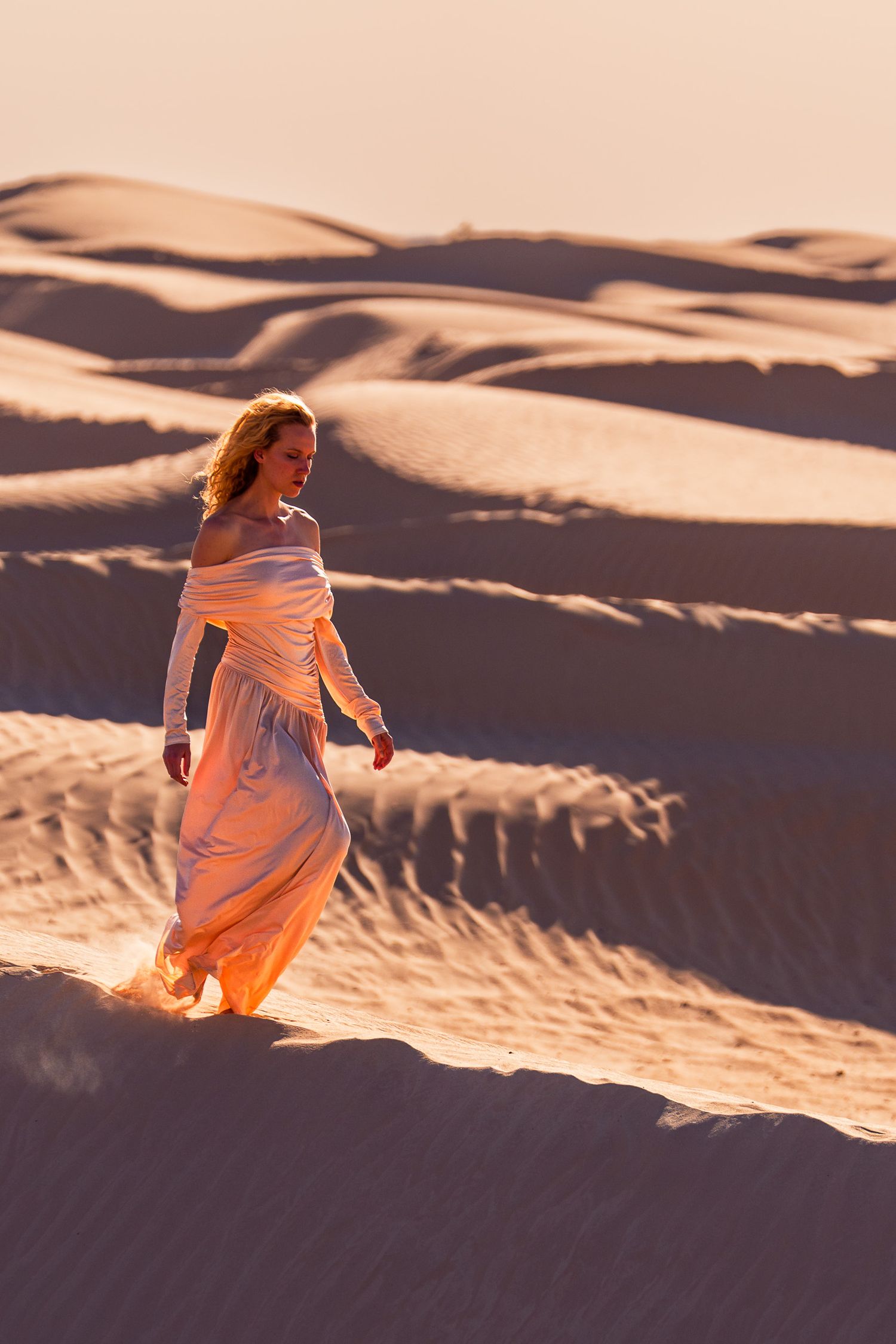 Woman in flowing off-shoulder gown walks gracefully across golden sand dunes at sunset.