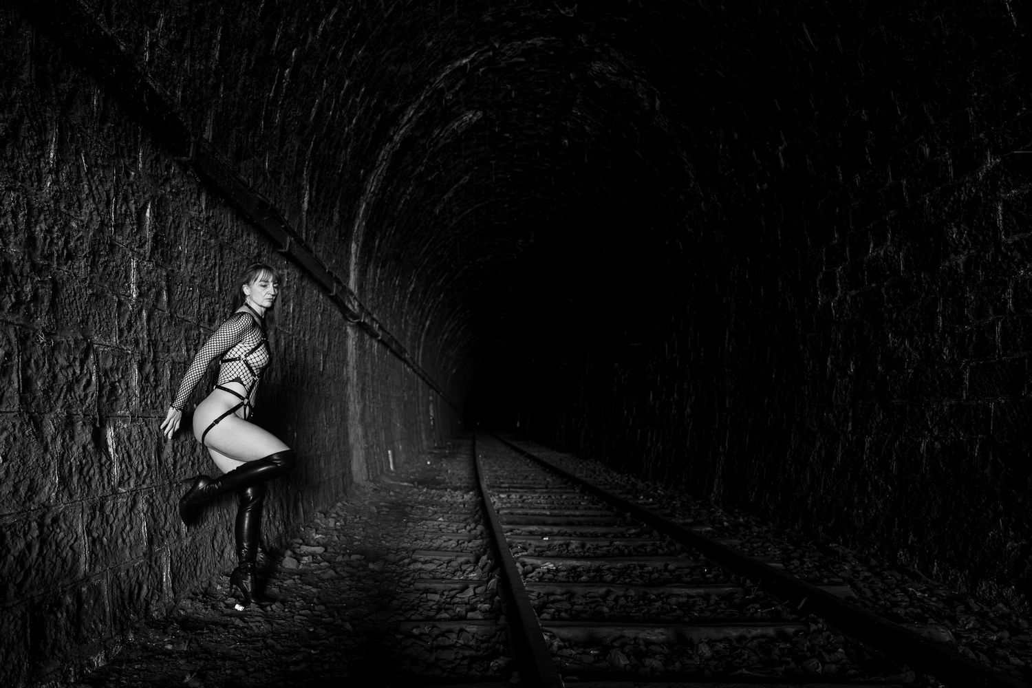 Woman in dark tunnel posing beside railway tracks in black and white dramatic photography.