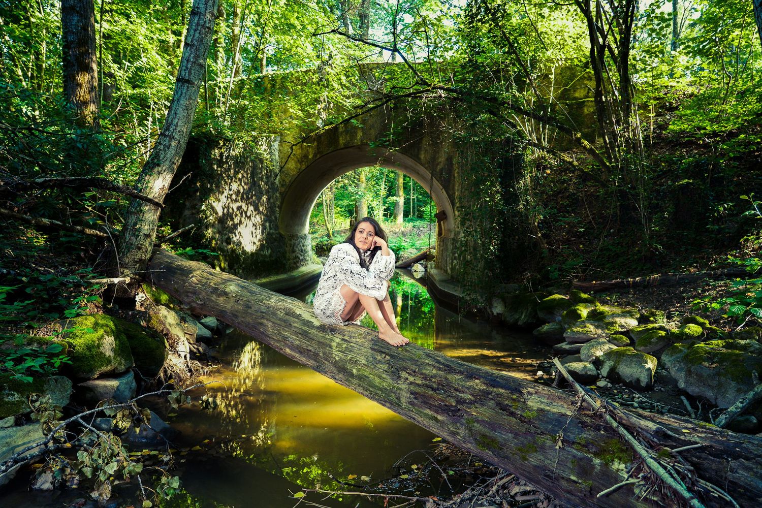 Woman sitting on fallen log by a mossy stone arch bridge over a tranquil forest stream surrounded by lush greenery.