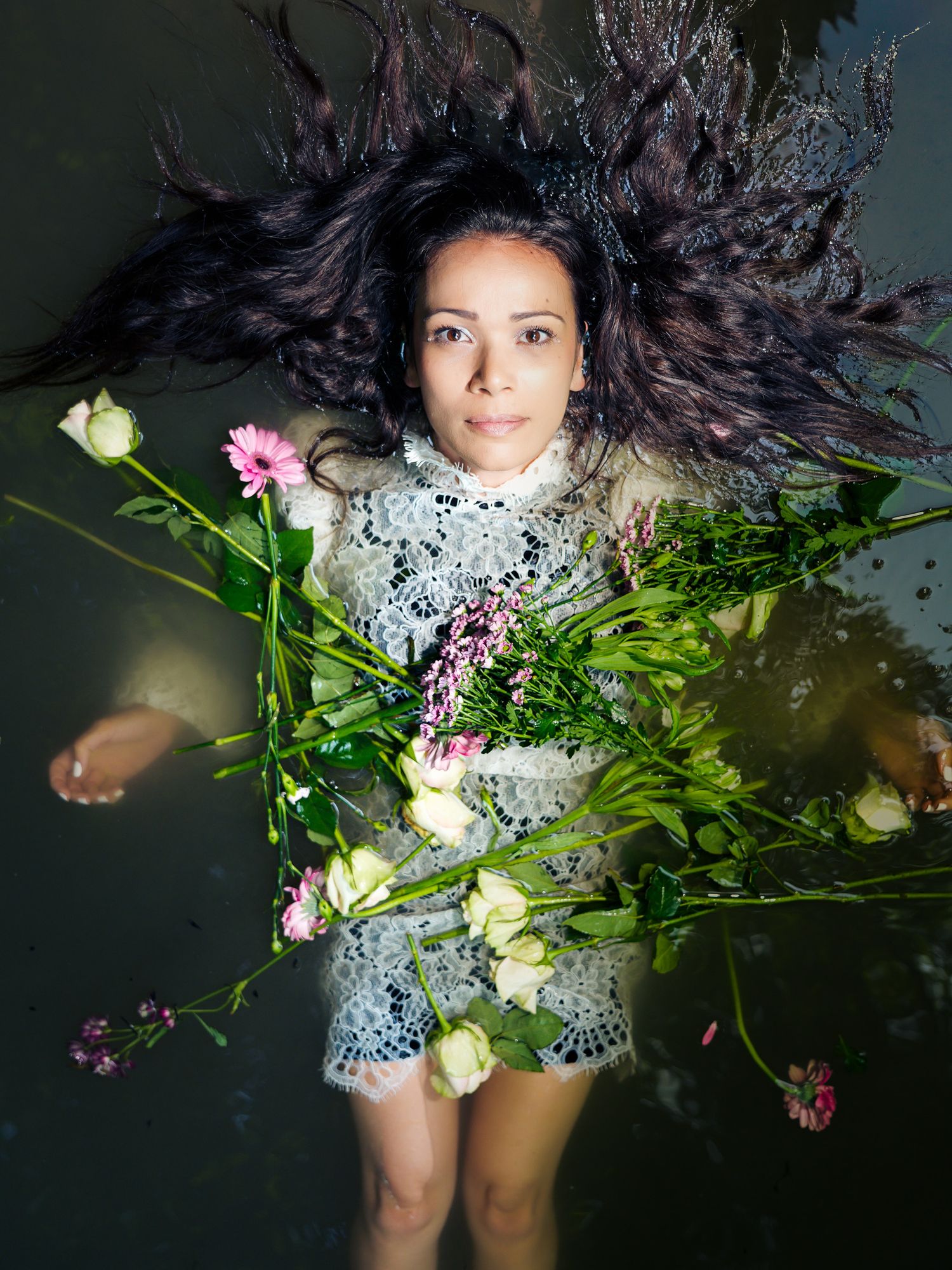 Woman floating in water wearing lace dress, surrounded by pink and white flowers with hair spread out.