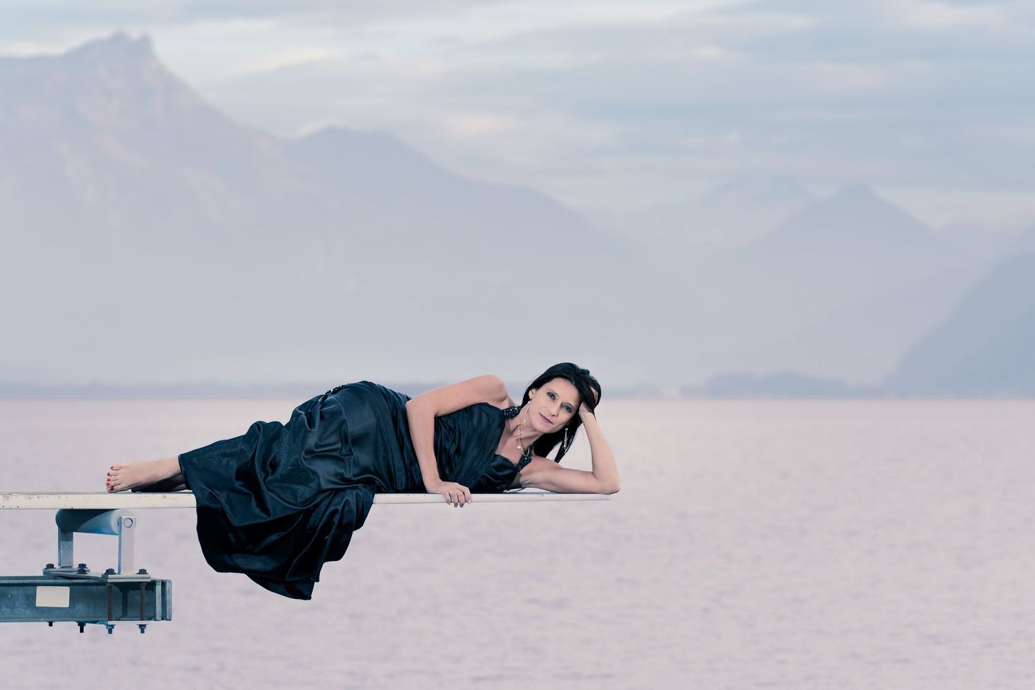 Woman in black dress lying on a surface with mountain backdrop in a serene, misty landscape.