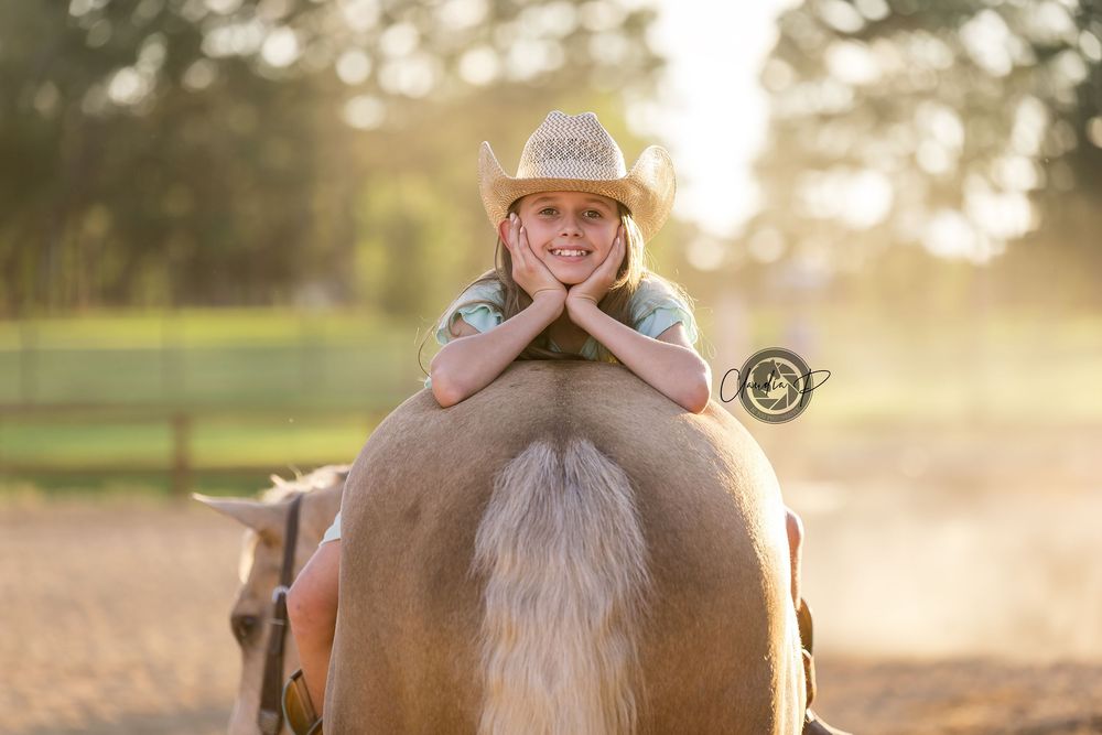 Smiling young girl in cowboy hat resting chin on hands while sitting on palomino horse.
