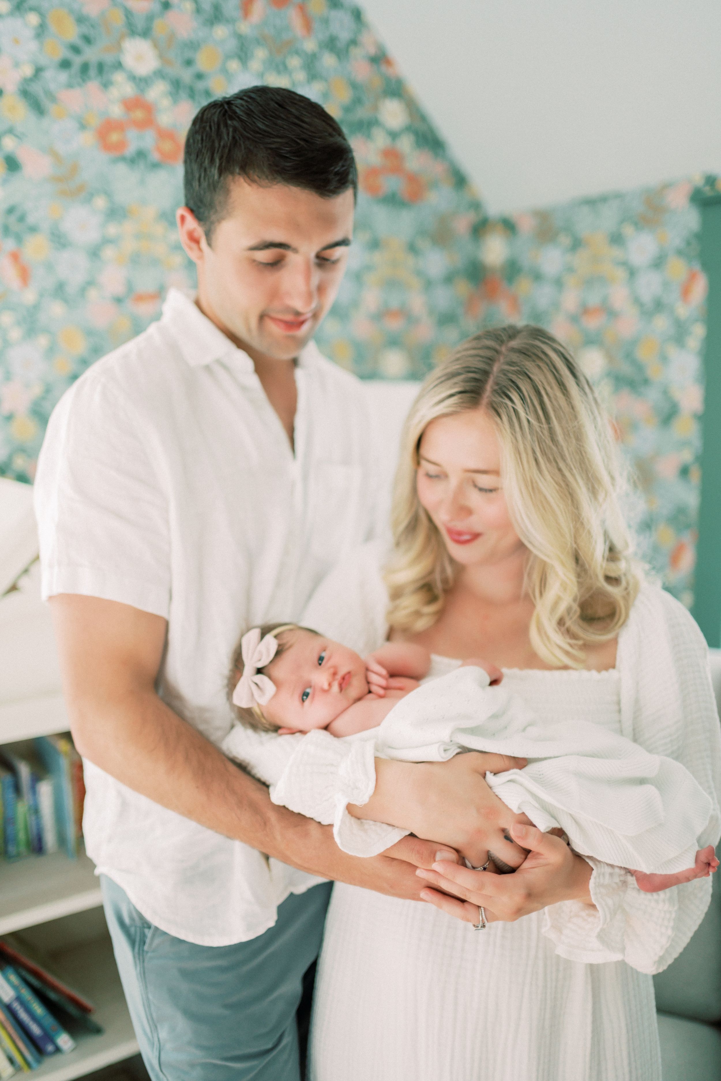 Parents in white outfits pose with their newborn baby in four tender nursery portraits with floral wallpaper background.