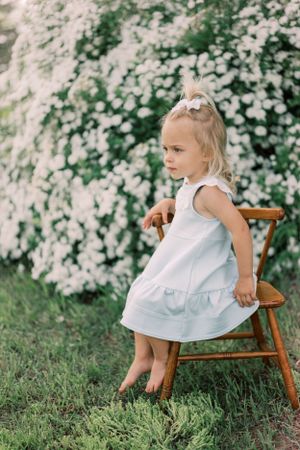 Small child in white summer dress standing against wooden chair with blooming floral backdrop in garden.