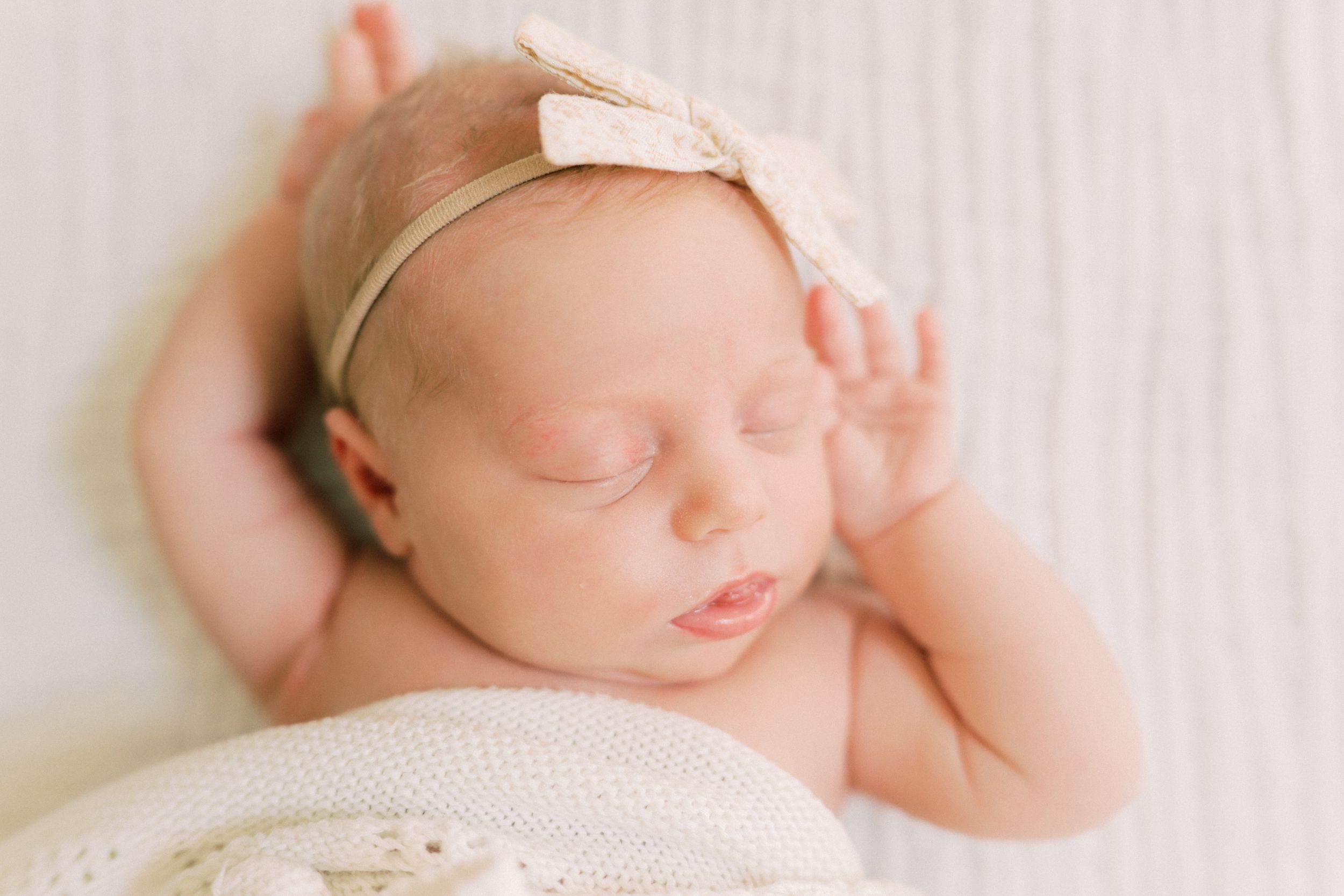 Peaceful sleeping newborn baby wearing delicate bow headband rests on soft white blanket.