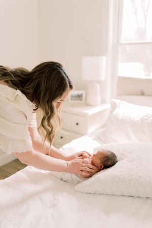 A tender moment captured in a bright bedroom as a woman in white gently attends to a newborn baby on white bedding.