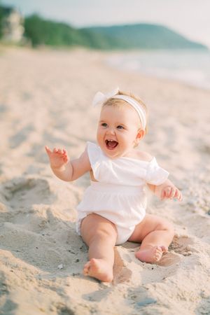 A happy baby wearing a white outfit sits and plays on a sandy beach with mountains visible in the background.