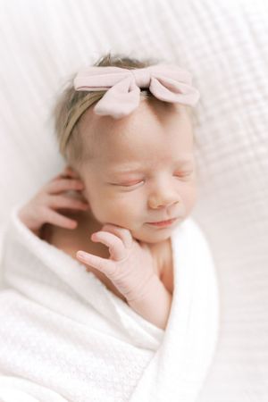 A sleeping newborn baby wrapped in white swaddle blanket wears a light pink bow headband while resting peacefully.