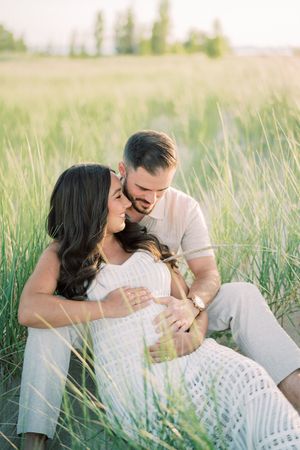 A couple in light colored clothing shares tender moments while sitting together in a field of tall grass at sunset.