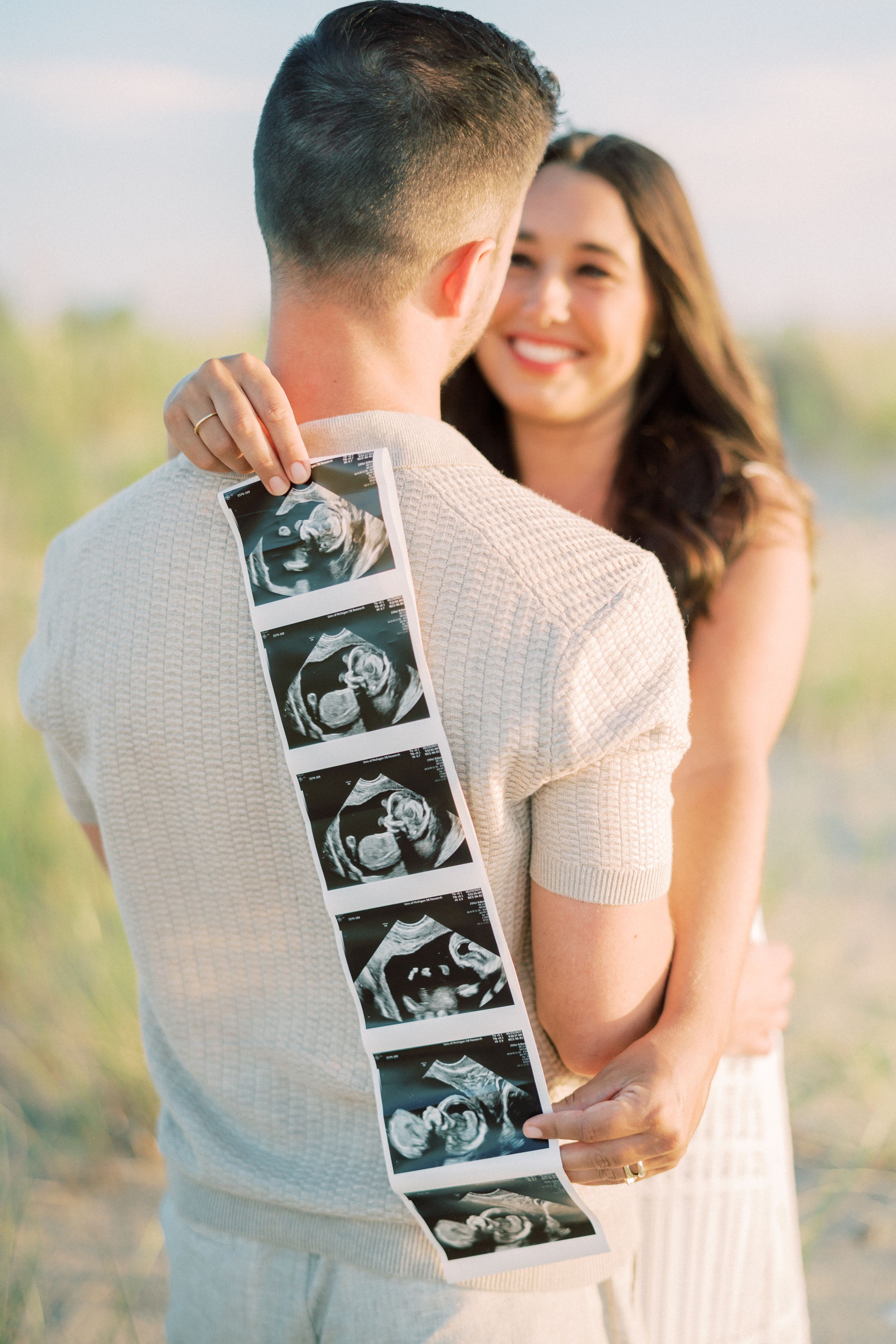 Couple embraces while holding up ultrasound photos in series of romantic pregnancy announcement pictures outdoors.