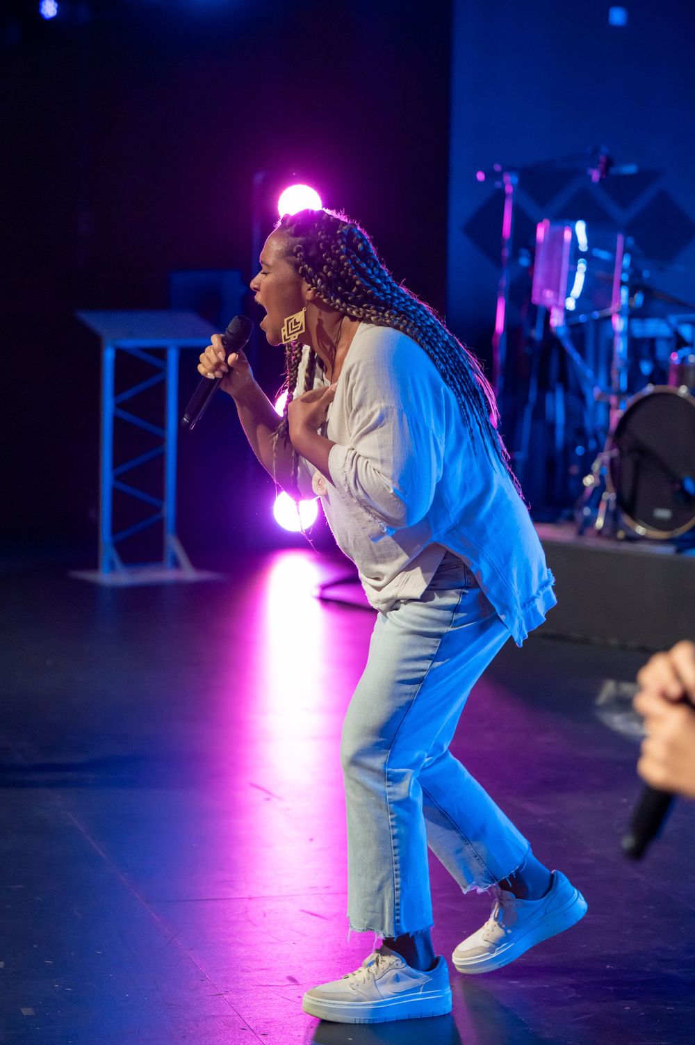 A singer with long braids performs energetically on stage under pink and blue spotlights.