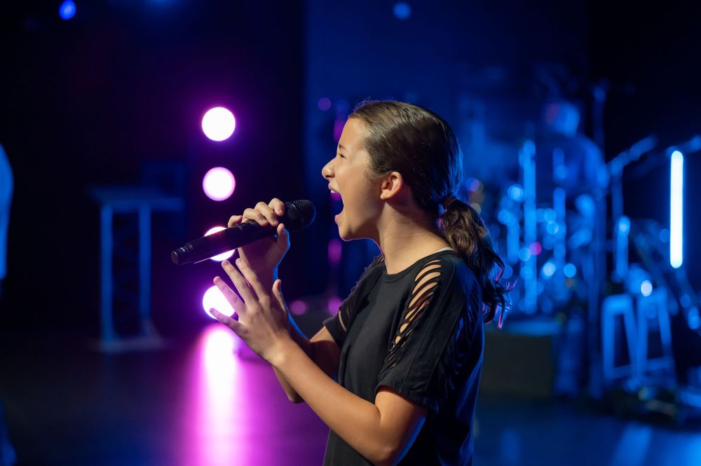 A performer sings passionately into a microphone on a dark stage with purple lighting.
