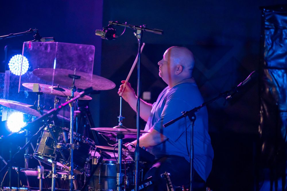 A drummer performs behind a drum kit on stage with blue stage lighting and effects.