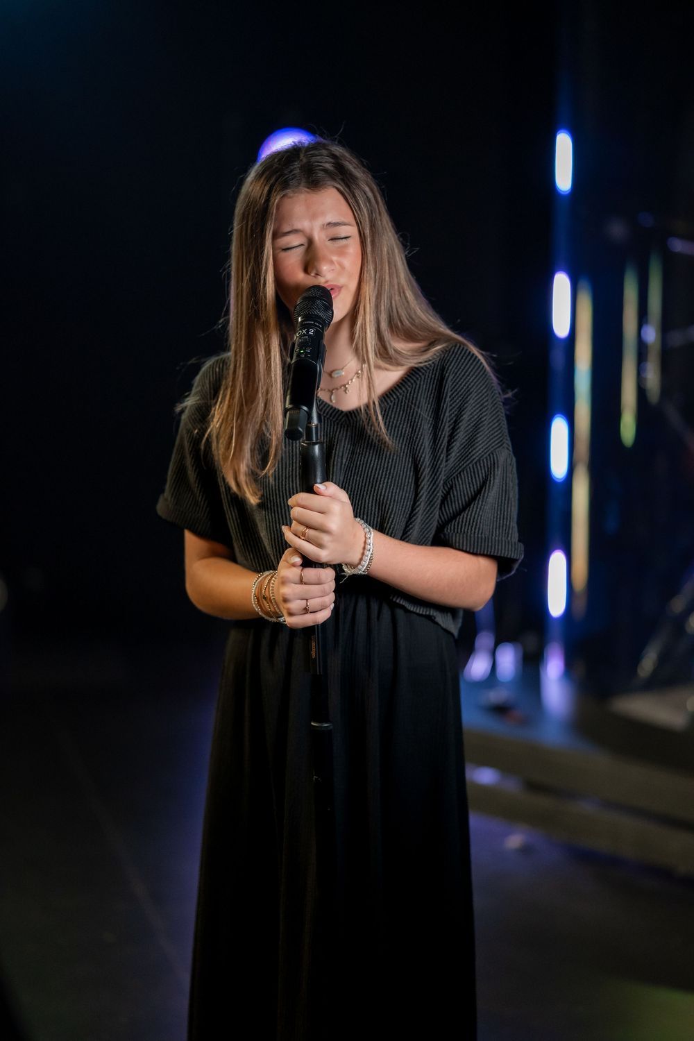 A person in a dark outfit sings passionately into a microphone against a dimly lit background with blue lights.
