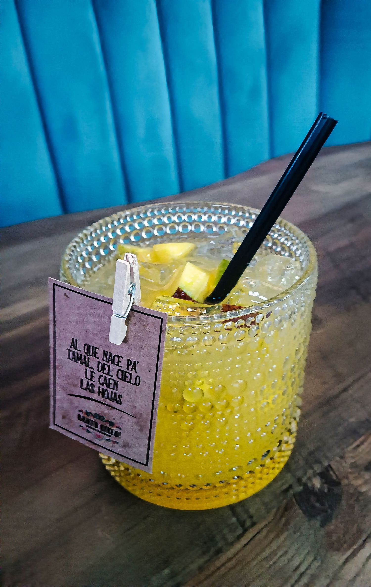 Yellow cocktail in textured glass with lemon garnish and drink tag against blue background.
