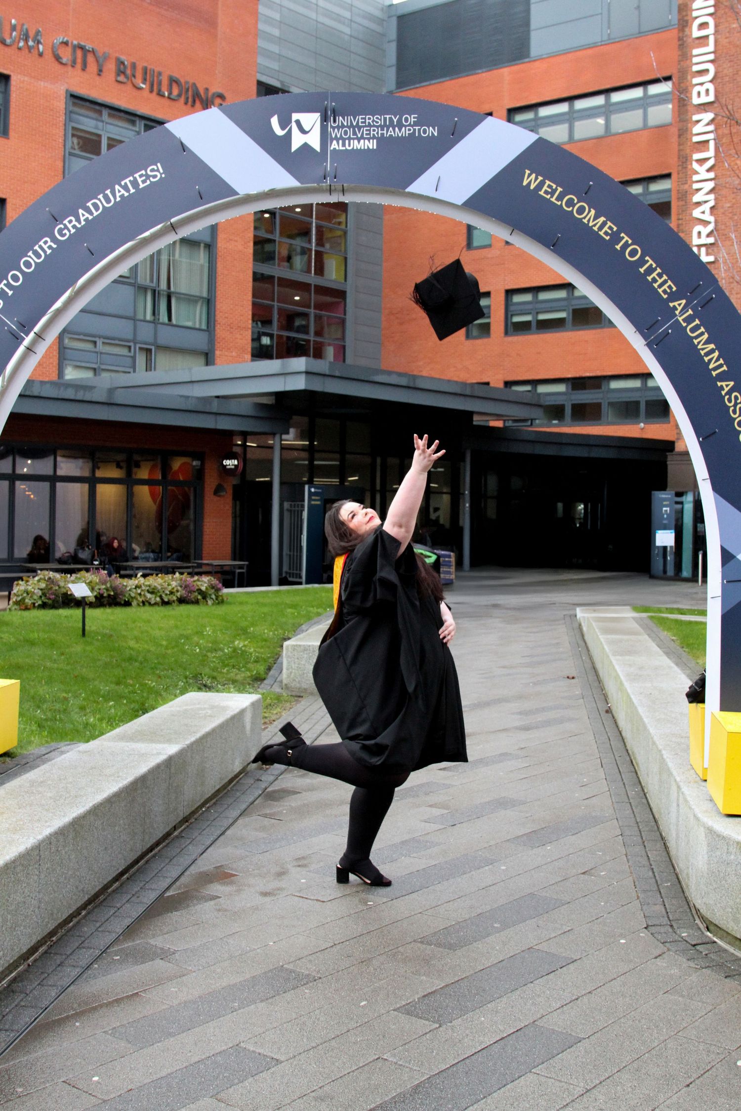 Graduate in cap and gown poses playfully by concrete steps near university building.