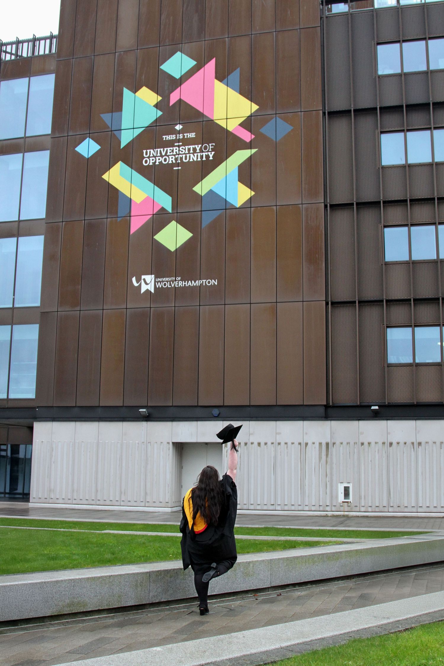 Graduate in cap and gown poses with colorful geometric university logo on modern building exterior.