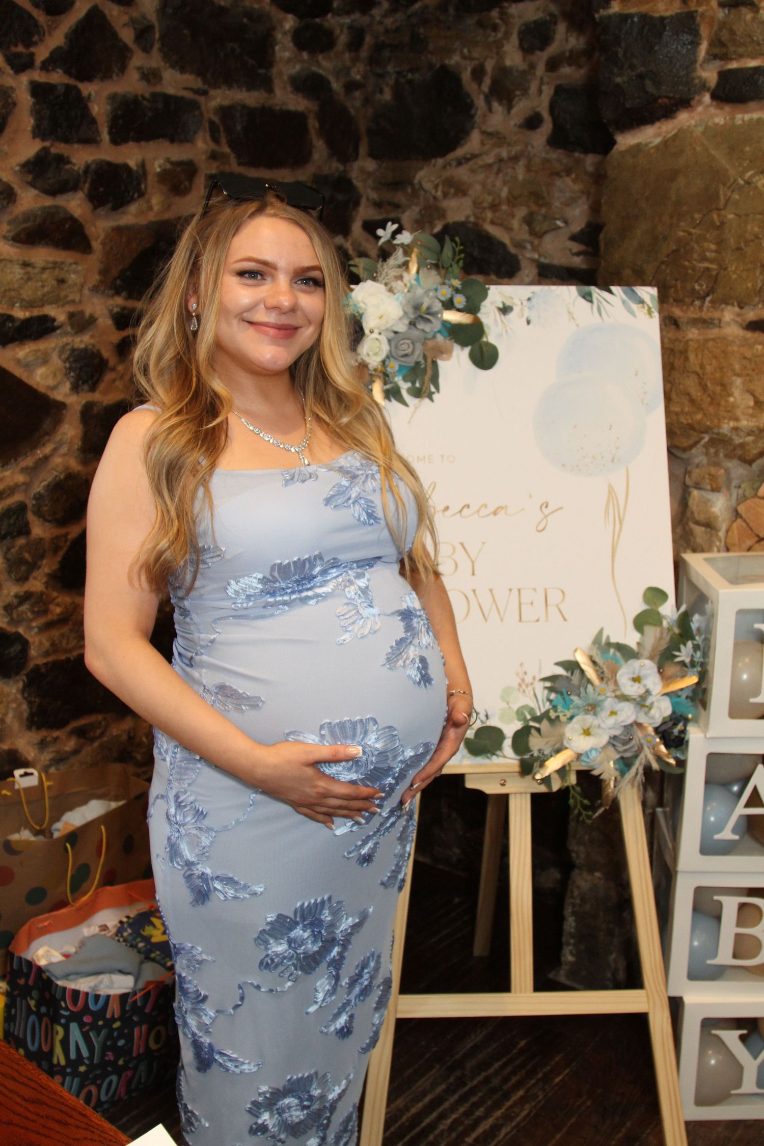 A light blue floral maternity dress against a rustic stone wall backdrop.