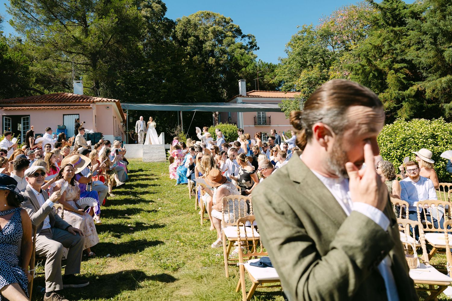 Outdoor wedding ceremony with guests seated on chairs in a sunny garden beside a pink building. Bride entering with father while groom cries