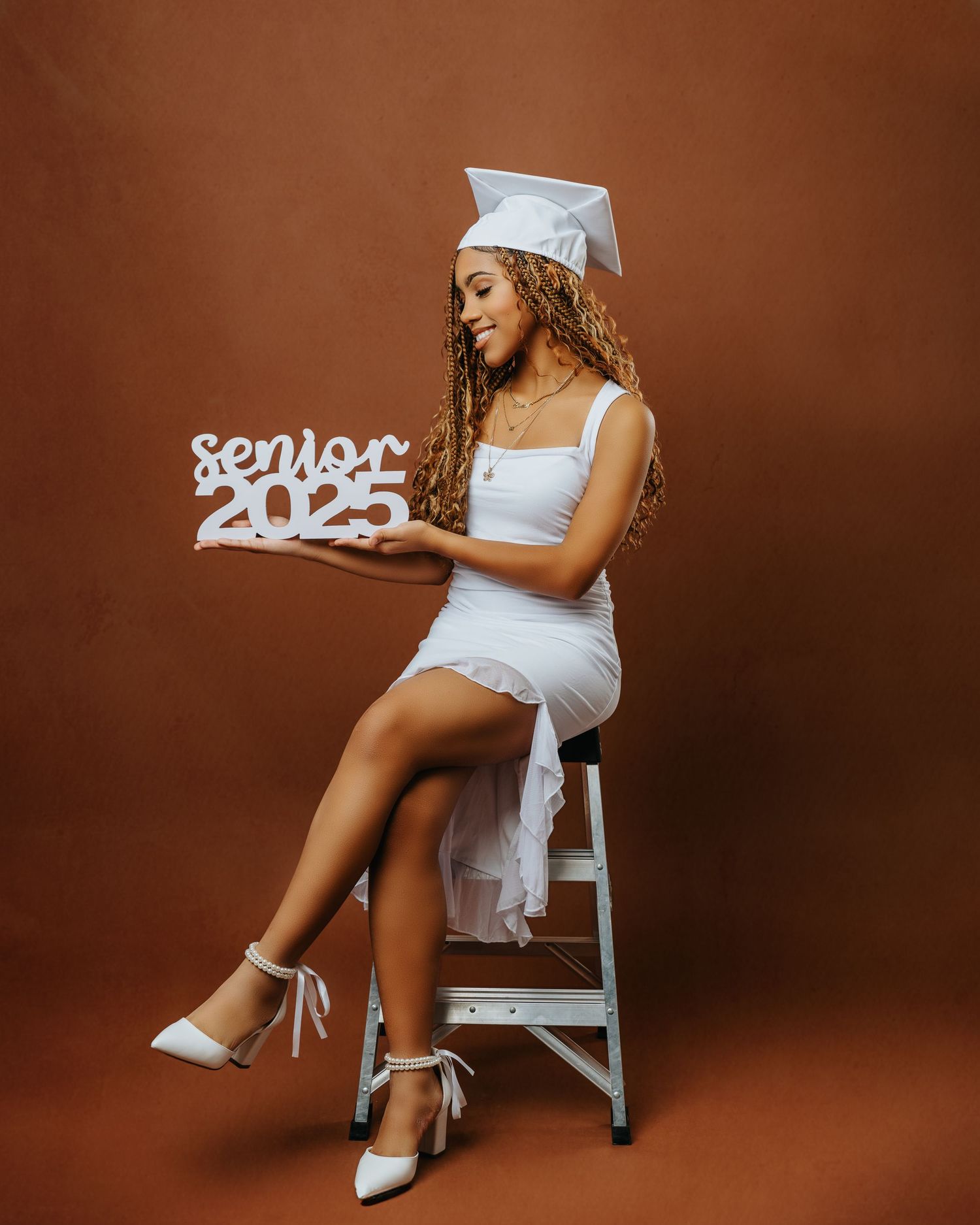 Graduate in white dress and cap poses with Senior 2023 sign on brown background.