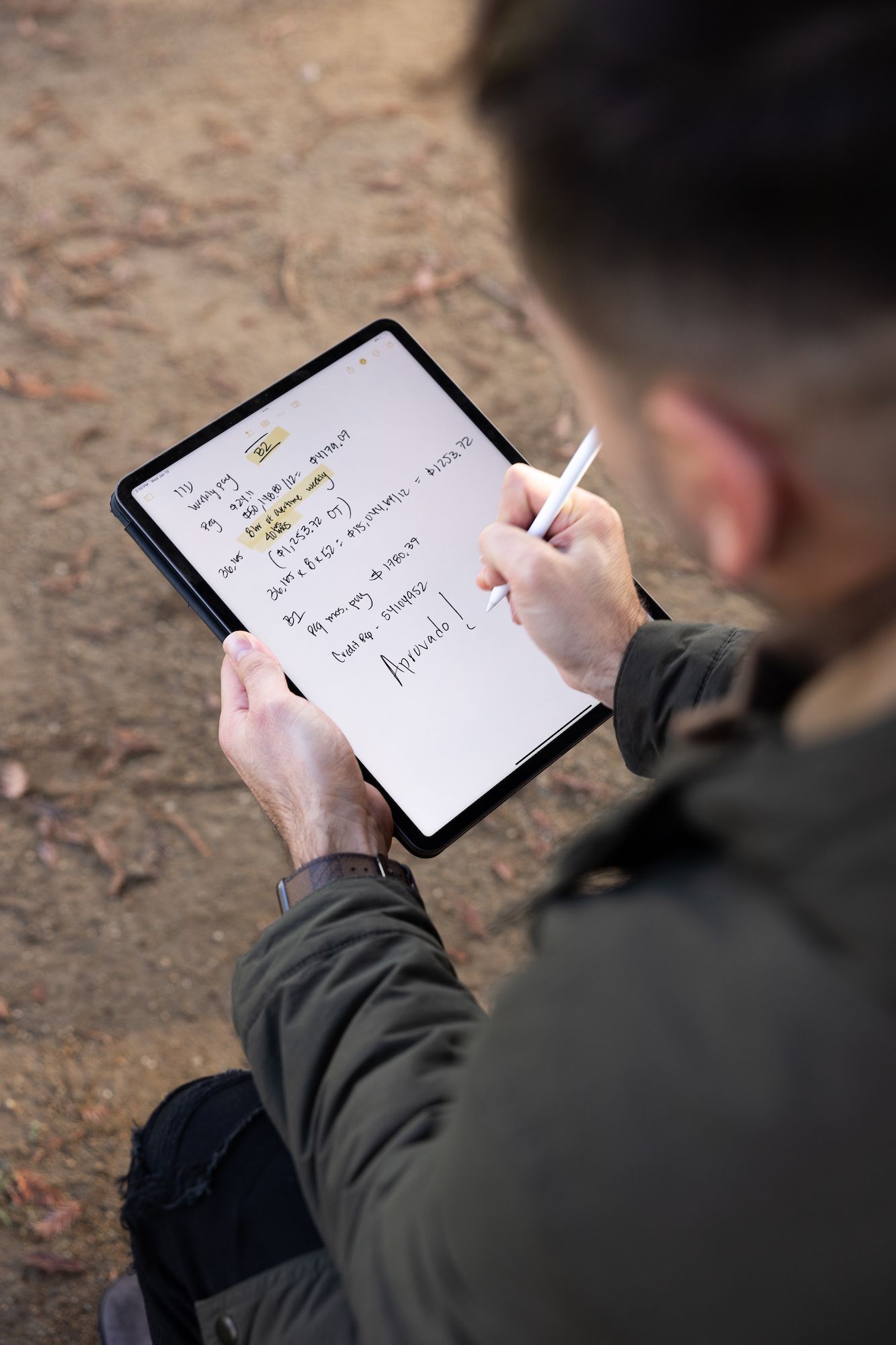 A man writes notes on a tablet with a stylus while sitting outdoors in a green jacket.