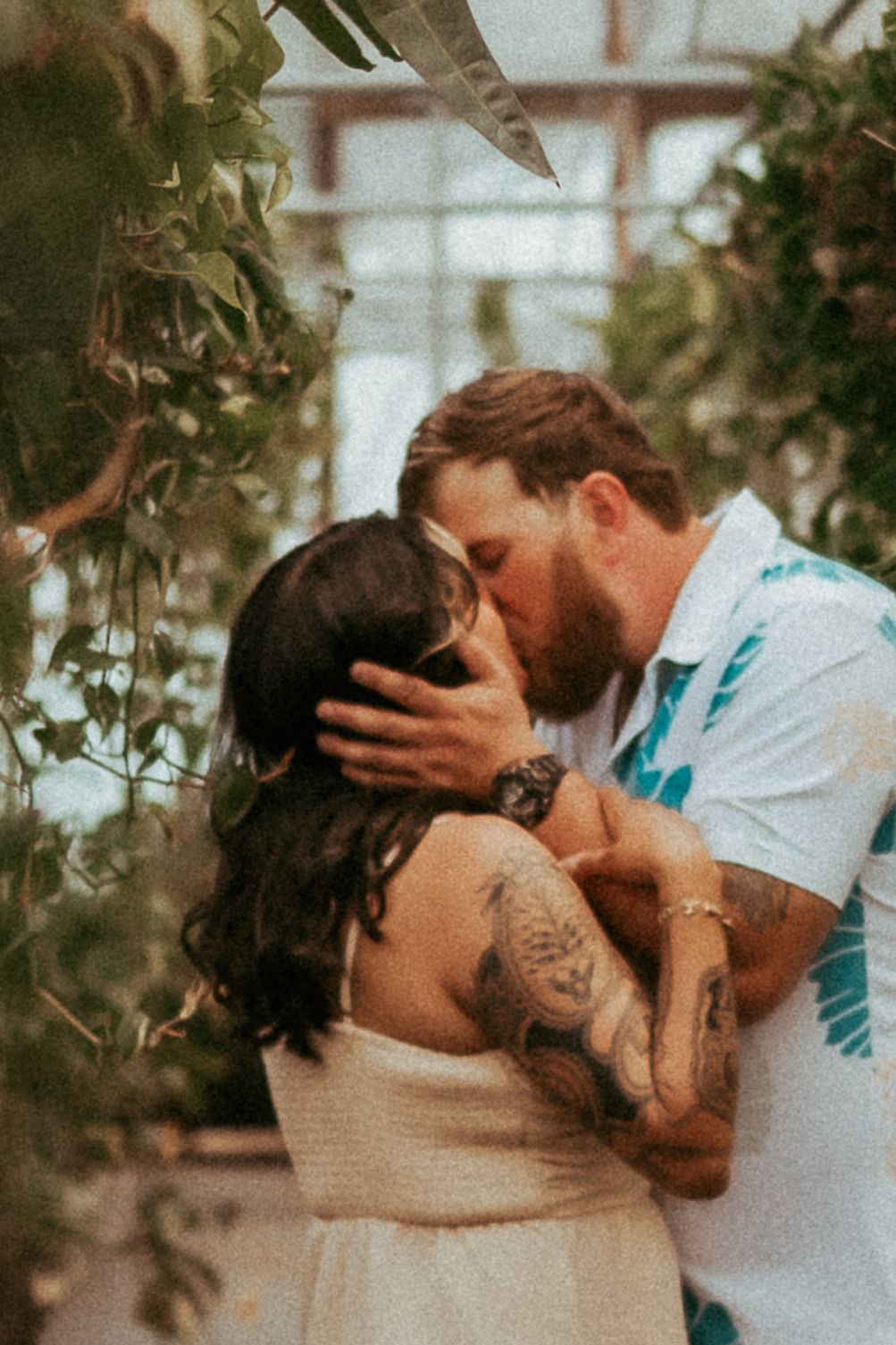 A romantic couple sharing an intimate kiss in a greenhouse with plants surrounding them.