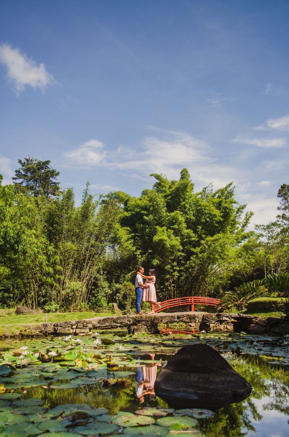 Couple embracing on a red arched bridge over a lily pond in a lush Japanese garden under a blue sky.