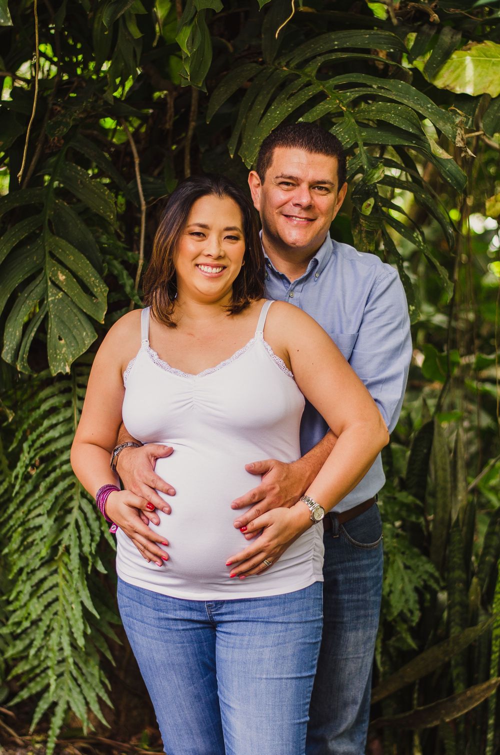 Happy pregnant couple posing outdoors among tropical green foliage, woman in white tank top cradling baby bump with partner behind her.